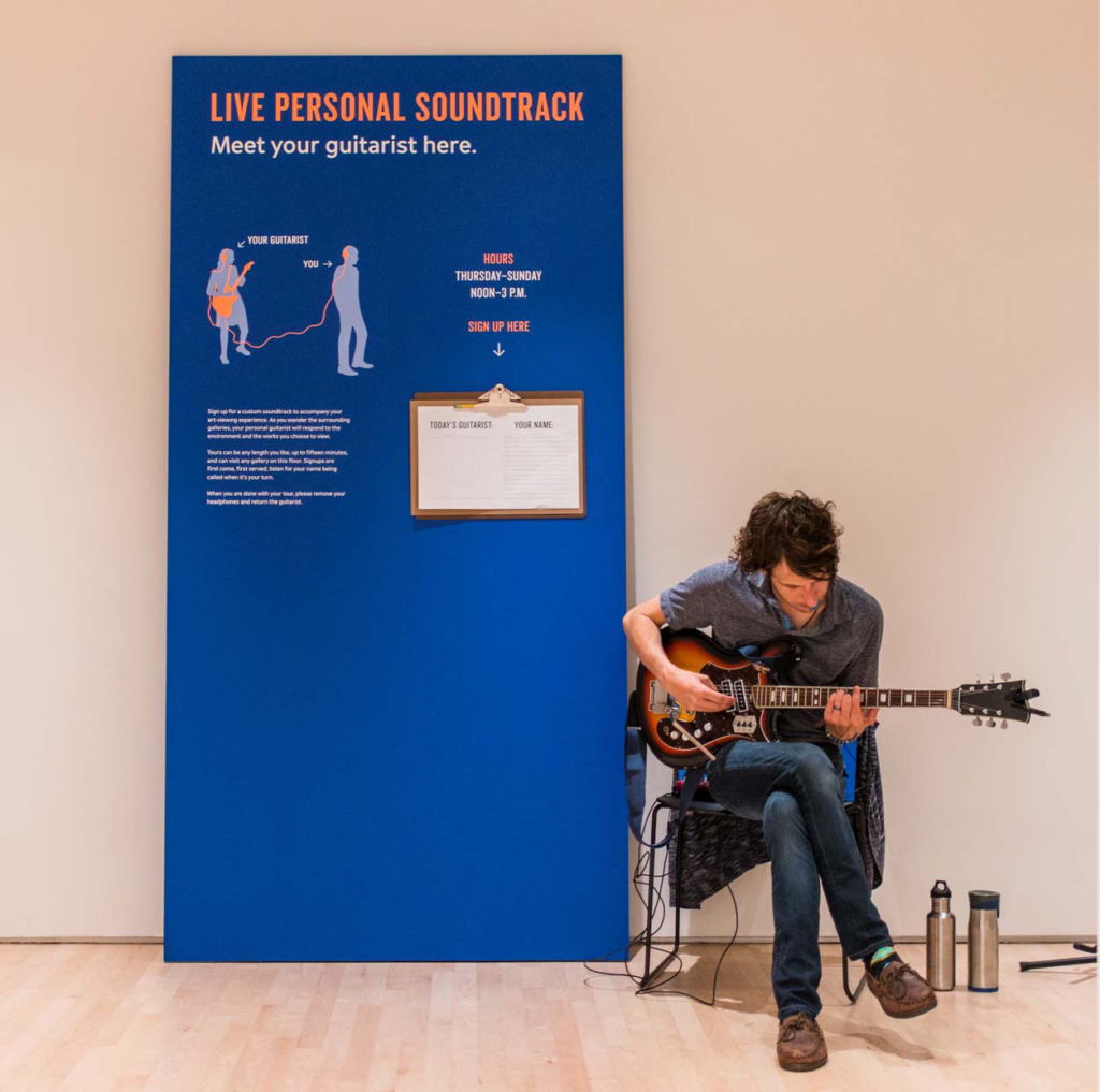 A Caucasian man sits holding a guitar in front of a sign that says "personal soundtracks", Kallmyer/Allen, Soundtracks