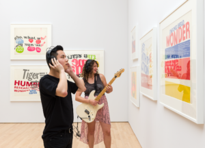 A Caucasian woman plays guitar while an Asian man listens wearing headphones in a gallery, Kallmyer/Allen, Soundtracks