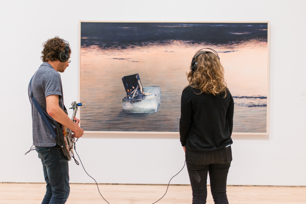 A man playing guitar stands next to a woman listening on headphones in front of a painting, Kallmyer/Allen, Soundtracks