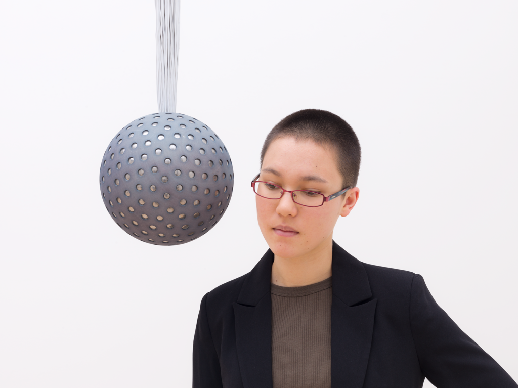 A woman wearing glasses with a shaved head leans into listen to a sphere suspended from the ceiling, Lozano-Hemmer, Soundtracks