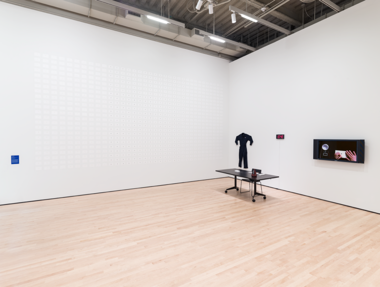 A gallery shows a grid of small white drawings, a jumpsuit, and a screen hanging on a wall before a desk, Munoz, Soundtrakcs