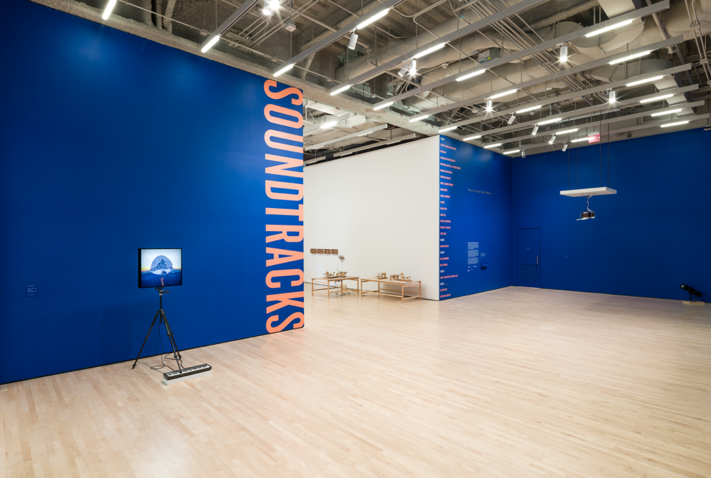 A video screen on a tripod before a blue wall with the words soundtracks written in orange vertically, Walker, Soundtracks