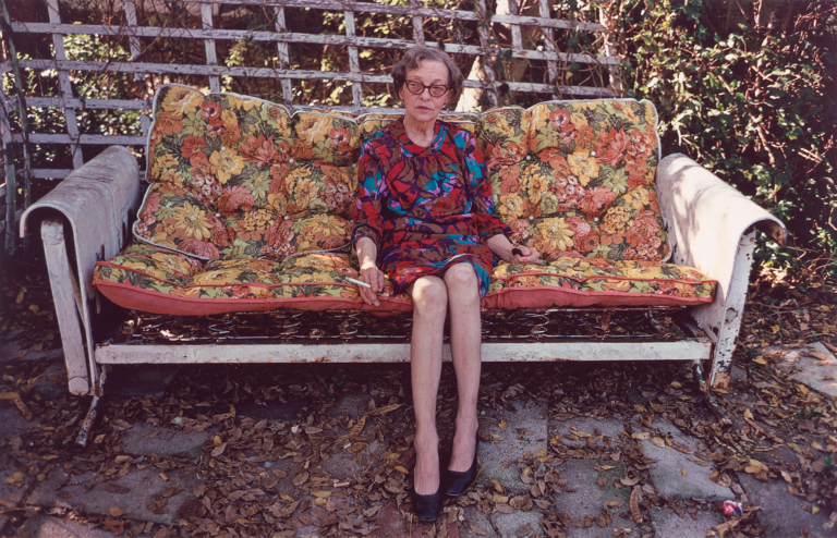 An elderly seated woman smoking a cigarette on a worn out floral print couch in an unkempt patio