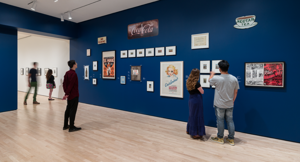 Visitors in a navy blue gallery looking at signs, photographs and other ephemera