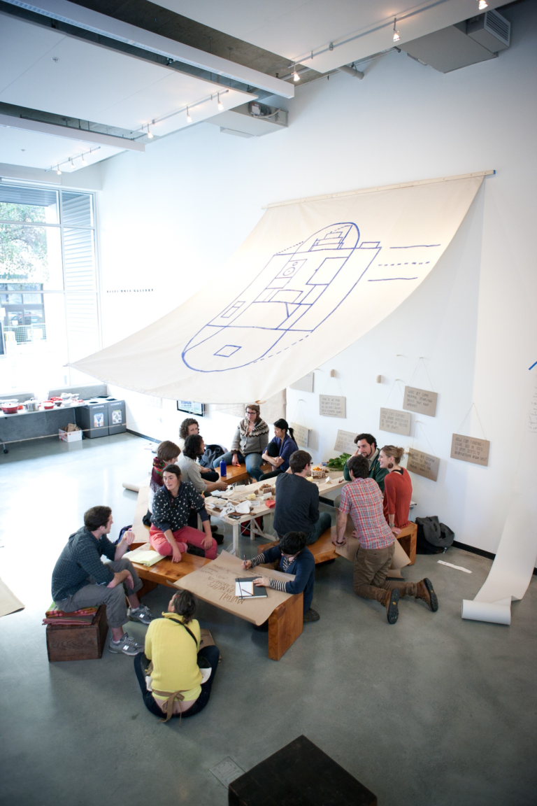 A group of young adults sit around an indoor picnic table with an canvas srchitectural floorplan hanging over them