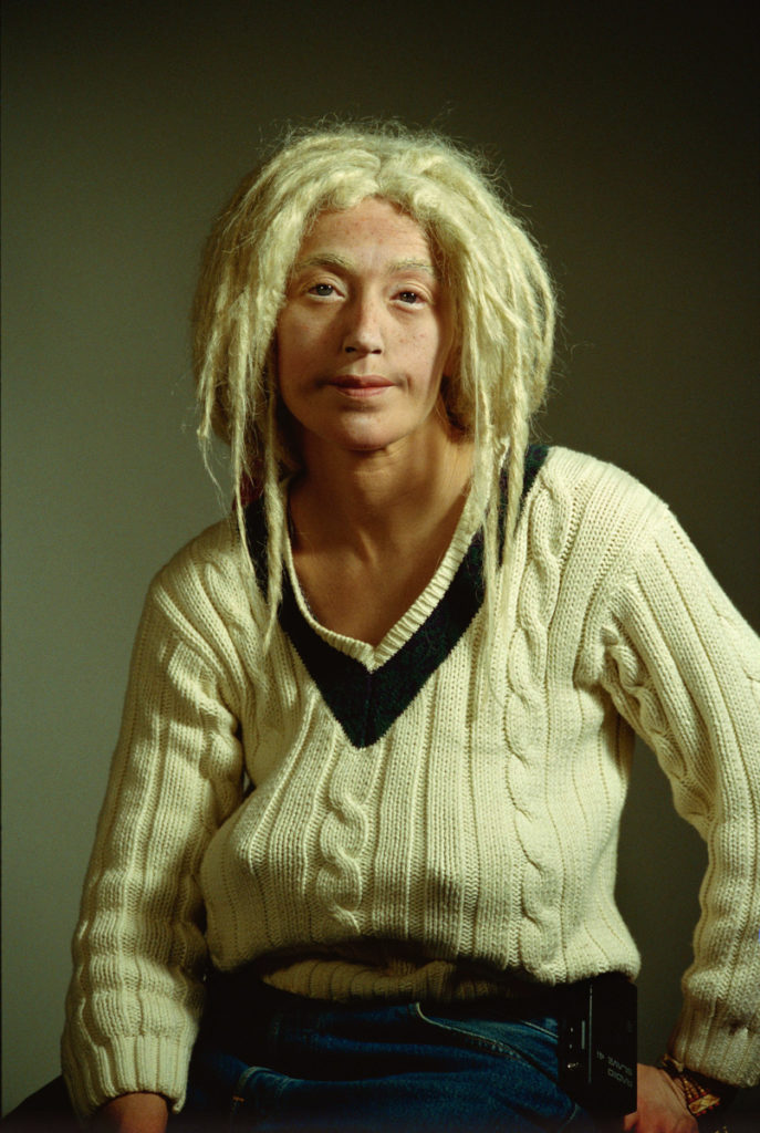 Studio-style color portrait of a woman with blonde dreadlocks and an ill-fitting, beige, cable knit sweater