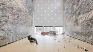 Artist Julie Mehretu sits back in a chair looking at two giant abstract paintings which take up two whole walls of her studio