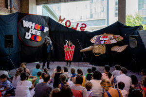 Performers dressed as a werewolf and a soda can on a stage with a large witchy puppet and a sign reading, "Whoop Dee Doo" behind