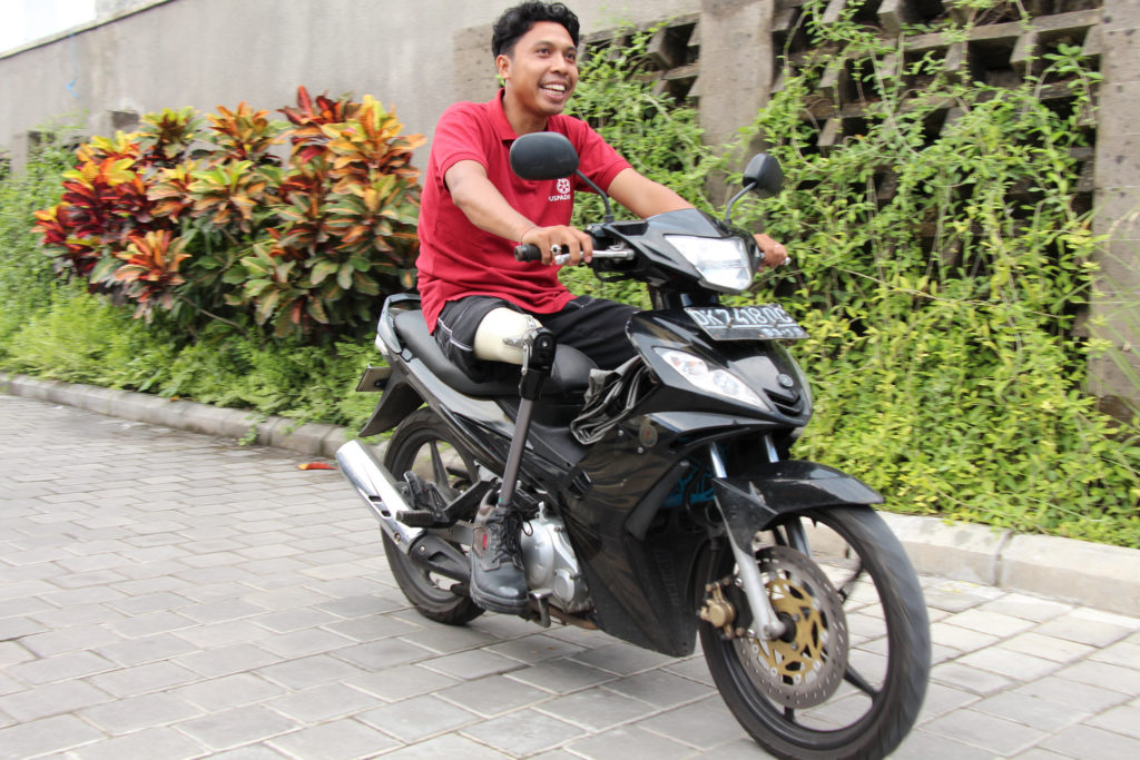 A man in a red t-shirt on a motorbike parked in a plant-lined paved driveway
