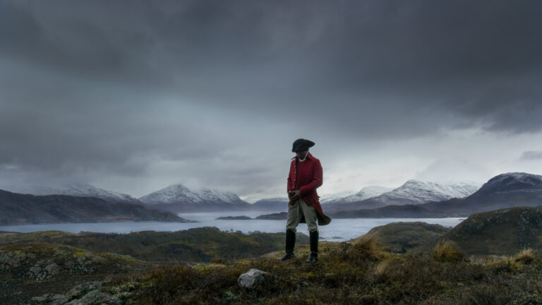 A black man dressed as an 18th century British sailor against a dramatic landscape