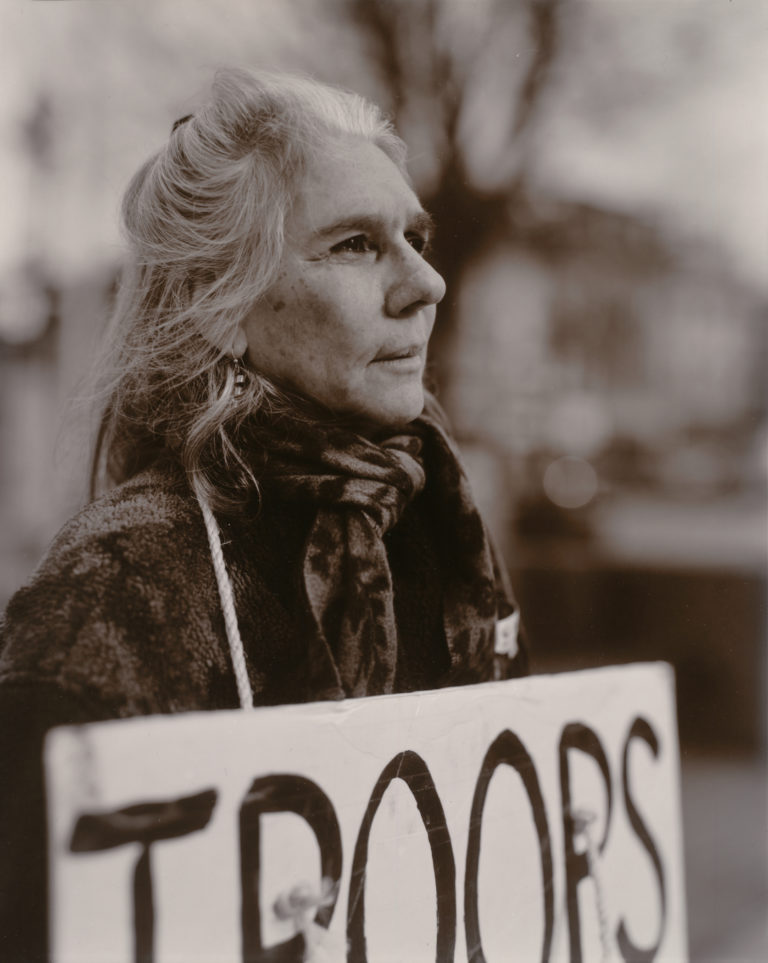 A black and white portrait of a woman with white hair holding a protest sign reading "Troops