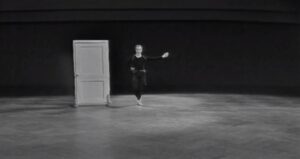 Black and white film still of a Caucasian man in black tights and leotard standing next to a door with one arm out streched, Antic Meet 
