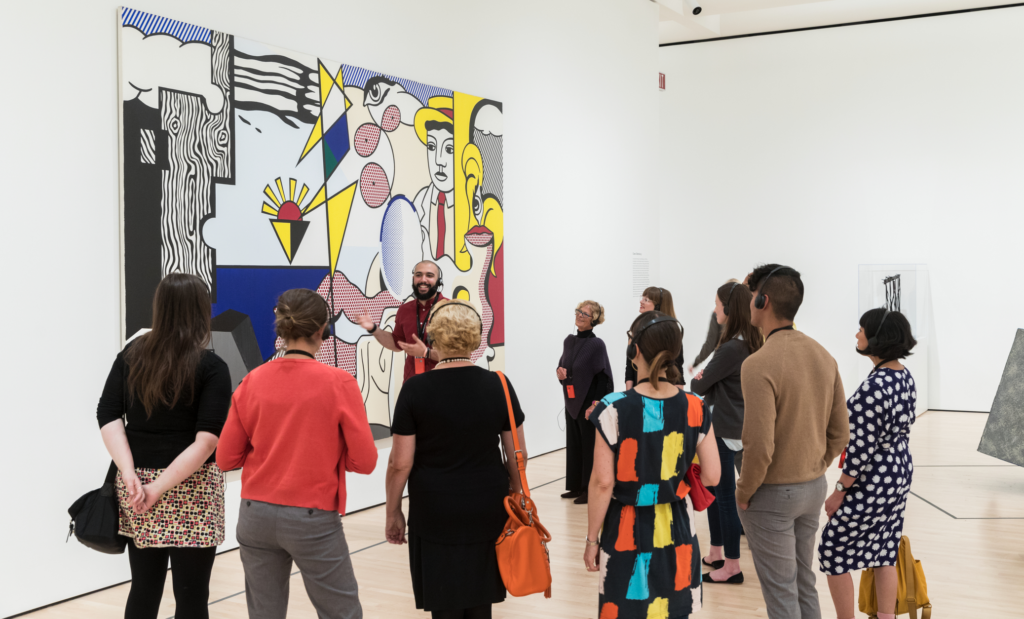 A group tour listens to a tour guide talk about a large painting by Roy Lichtenstein.