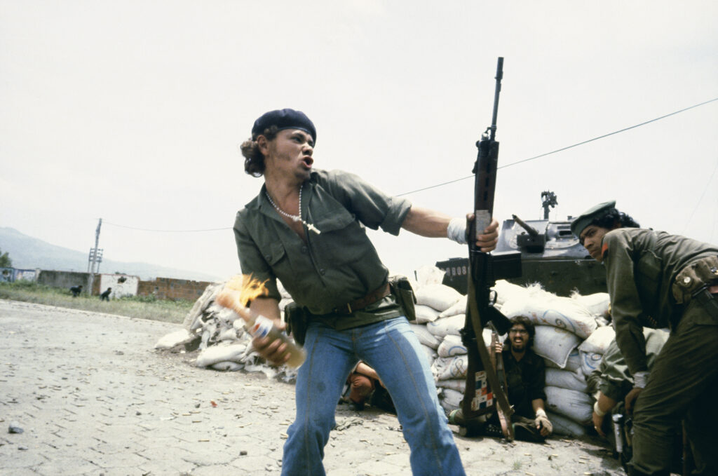 A man in a beret holding a rifle in one hand and throwing a Molotov cocktail made of a Pepsi bottle, with other men in uniform and a tank behind him