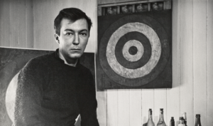 Black and white portrait of a young Caucasian man with brown hair sitting before a painting of a target, Rauschenberg