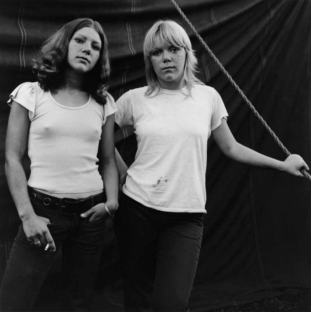 Black and white photo of two young women in tank tops and jeans standing in front of a curtain or tent