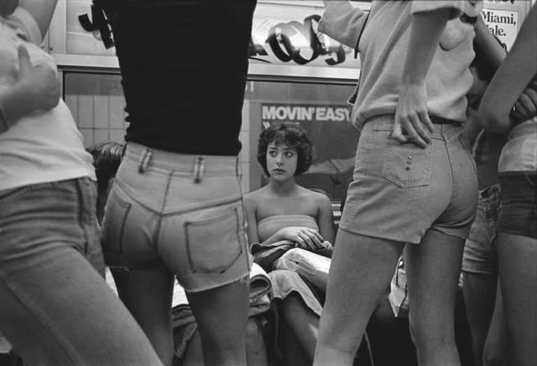 A seated young woman on a subway car with women in cut-off shorts standing in front of her