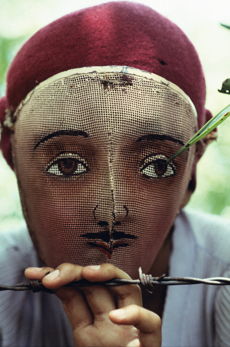 A closeup of a man or youth wearing a woven mask and burgundy beret and holding on to a barbed wire fence