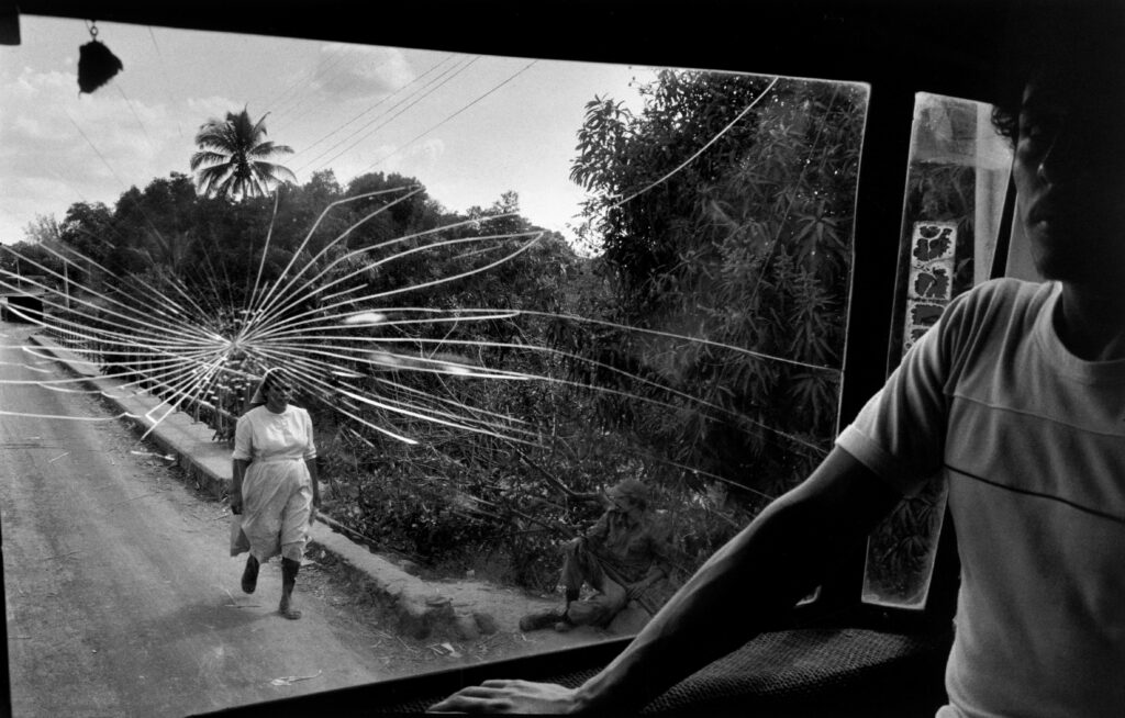 A man standing near the cracked window of a bus, which looks out onto a rural road where a woman in a white headscarf walks and a soldiers sits watching her