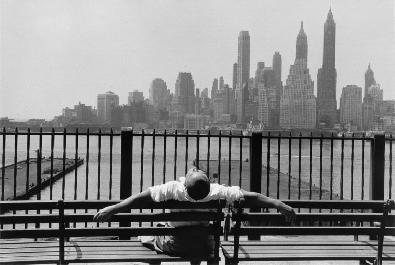 A man sitting on a bench with both of his arms behind it in front of manhattan skyline