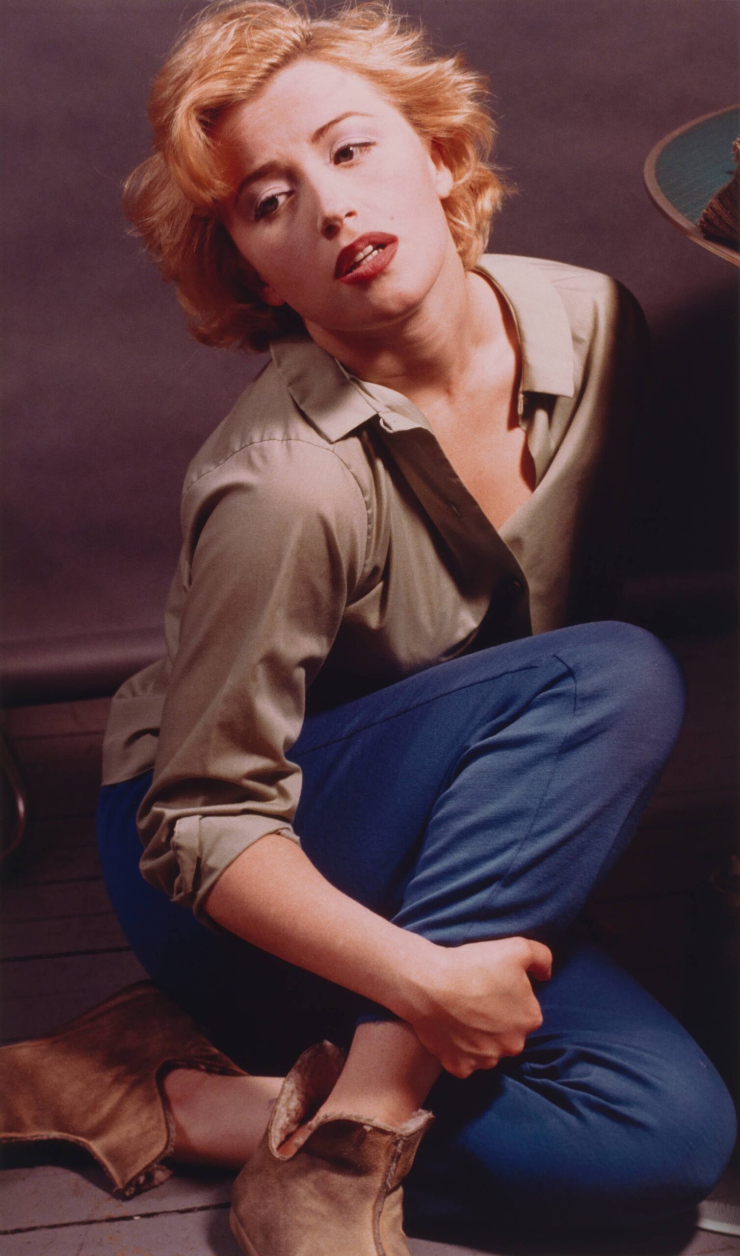 A photograph Cindy Sherman dressed as Marilyn Monroe wearing blue pants and a tan blouse and sitting on the floor.