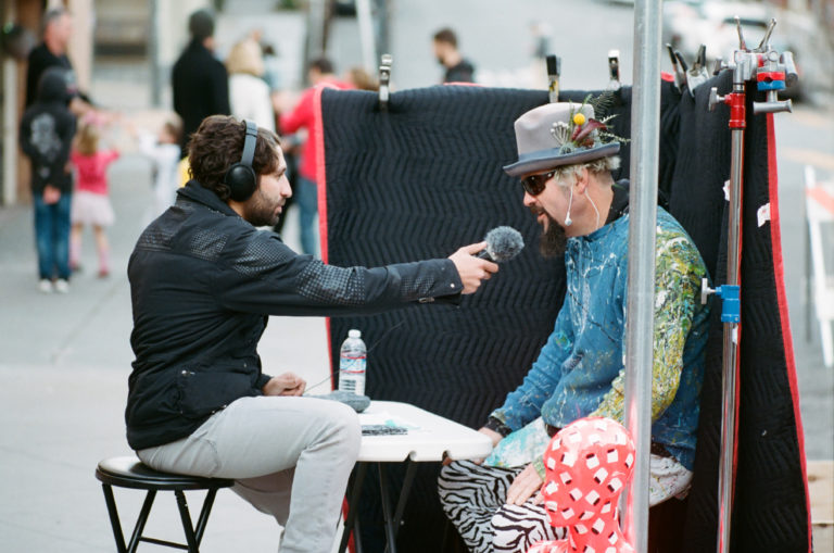 a person dressed in eccentric prints is interviewed on a sidewalk