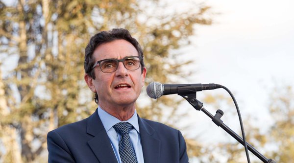 A man wearing a suit and glasses speaks at a podium with trees in the background