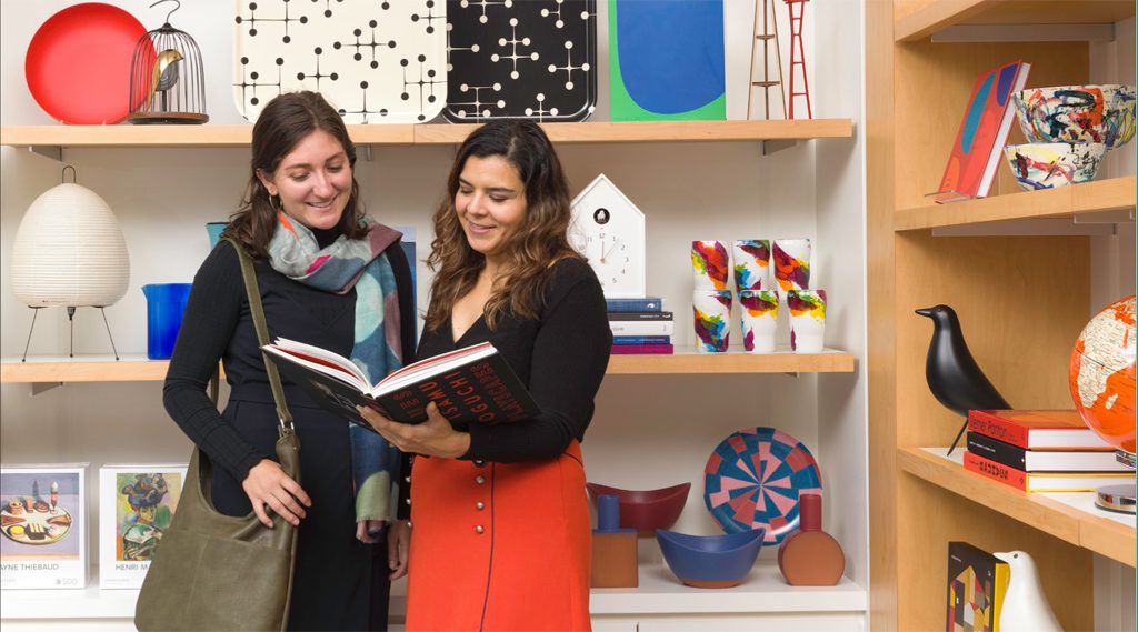 Two women smile while looking at a large book in the SFMOMA museum store with other products on the shelves behind them