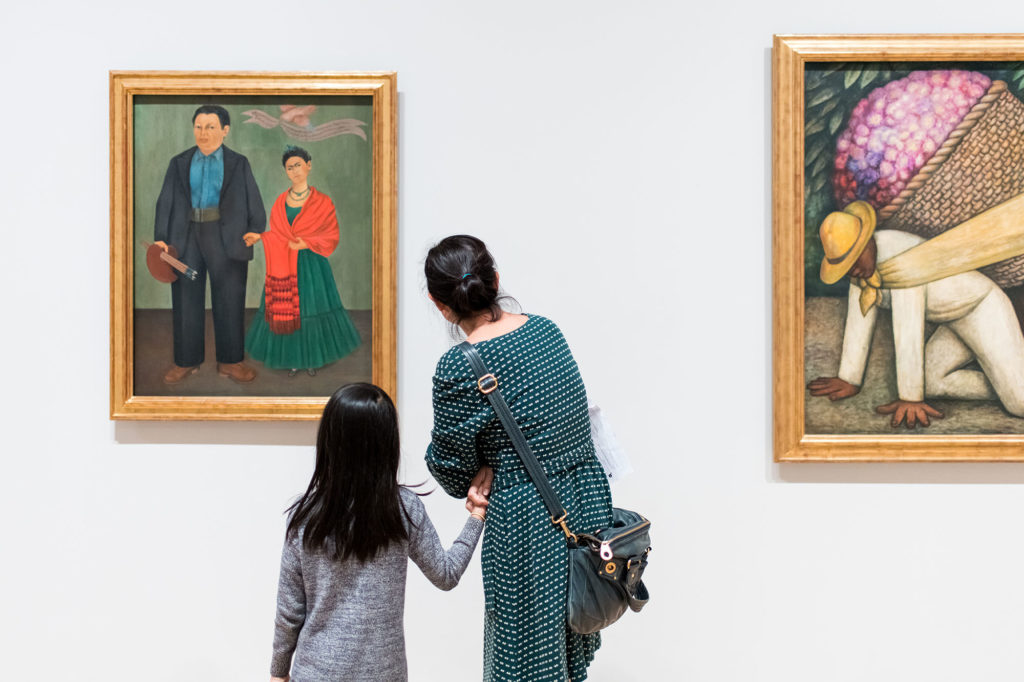 A parent and a child hold hands while looking at a painting by Frida Kahlo at SFMOMA.