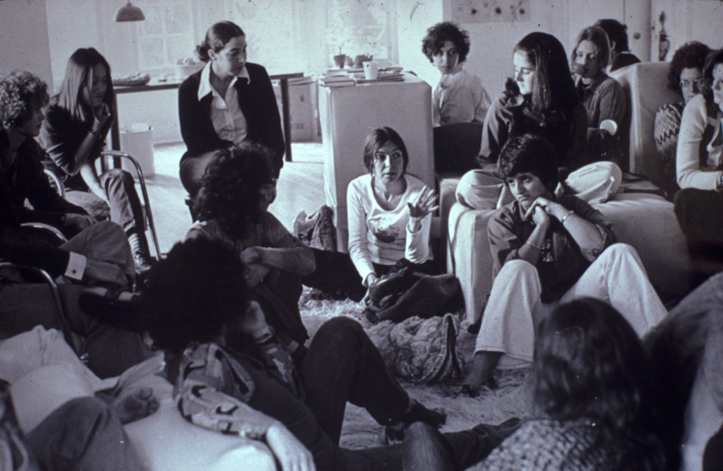 Black and white film still of a group of women wearing 1970's clothing sitting in a room having a discussion.