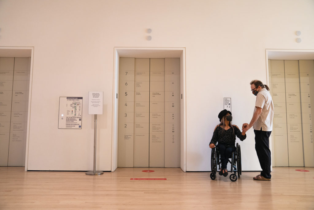 Two people, one standing and one in a wheelchair, hold hands as they wait for the SFMOMA elevators.