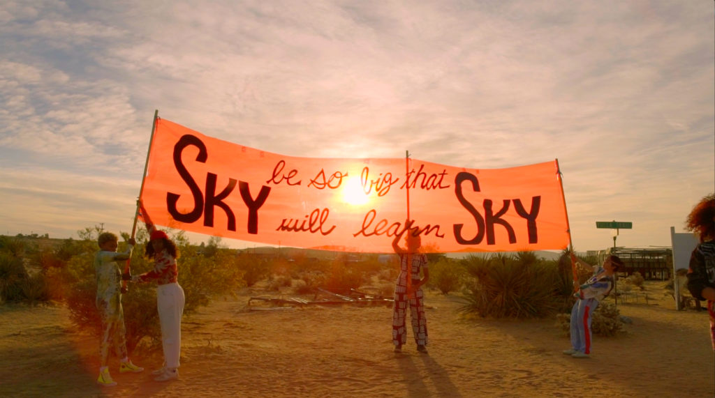 A film still depicting a desert landscape with four women holding a banner that reads, "be so big that Sky will learn Sky."