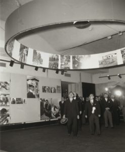 A black and white photograph of a group of Japanese men walking through an art gallery full of photographs.