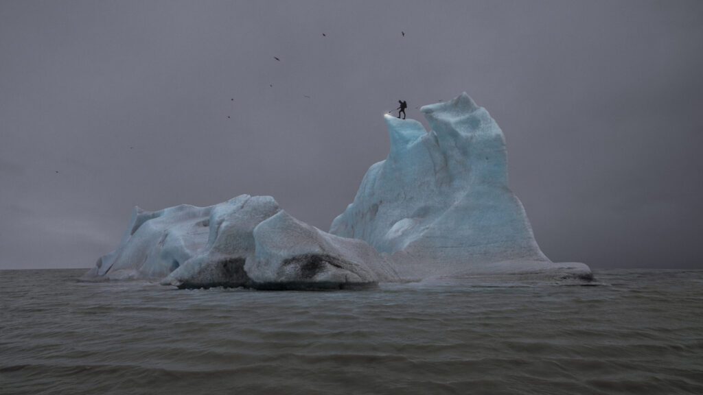 An artwork named The Blue Fossil Entropic III by artist Julian Charrière, depicts an arctic landscape capturing a person scaling a glacier at night.