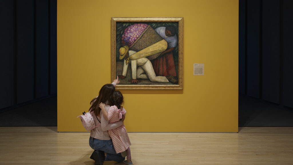 Woman and small child embrace while looking at "The Flower Carrier" (1935) by Diego Rivera