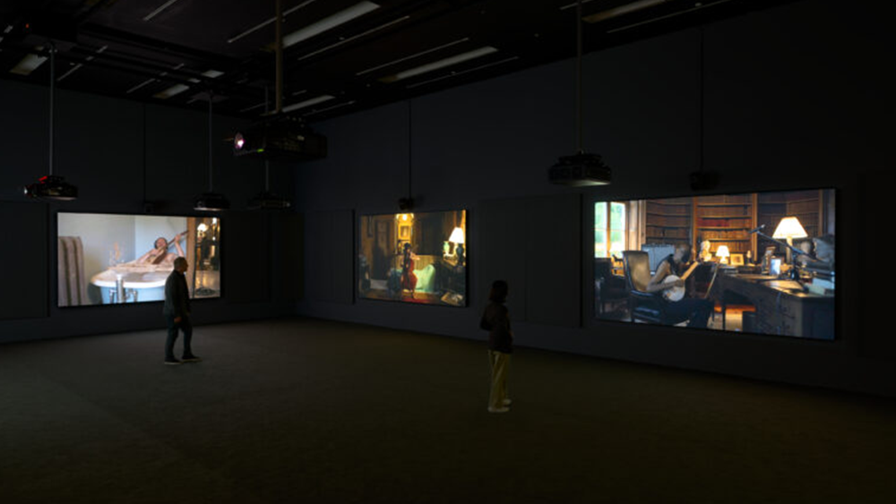 Two visitors stand inside an SFMOMA gallery in front of three out of nine screens showing Ragnar Kjartansson’s beloved video installation The Visitors.