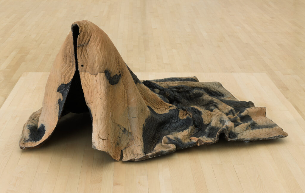 The artwork Tarpaulin No. 1, by artist Rebecca Belmore, is a ceramic blanket on the ground shaped as though draped over an invisible human figure.