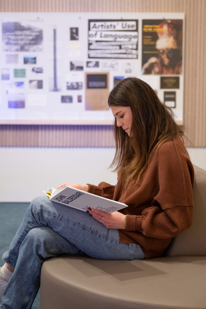 A woman wearing a brown sweater and jeans sitting on a cushioned seat in one of SFMOMA's learning lounges reading a book by John Baldessari.