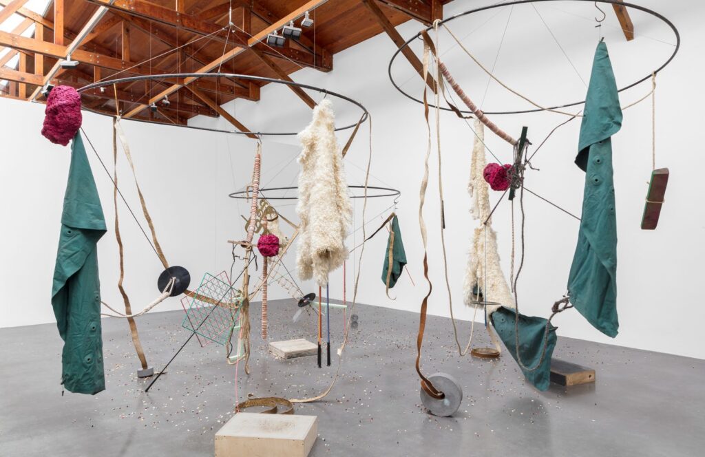 An abstract sculpture of three large metal hoops suspended from the ceiling with a variety of materials such as wood, fabric, lambskin, plastic, cardboard hanging from each hoop.