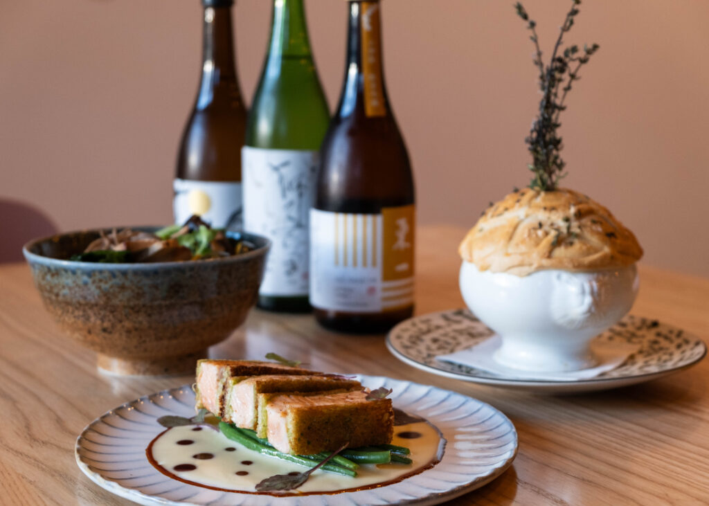 An artistically displayed assortment of food including chicken pot pie, salmon en brioche, soba noodles, and three bottles of wine in the background.