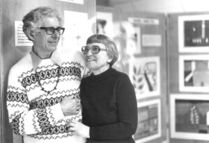 A black and white photograph of an elderly couple smiling and standing close together in what appears to be an exhibition or gallery space, with framed artworks visible in the background. The man is wearing a patterned sweater and glasses, and the woman is in a black turtleneck, also with glasses.