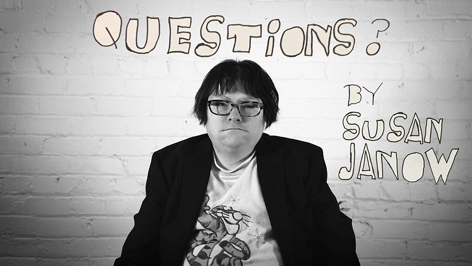 A black and white film still of a person sitting in front of a brick wall with the word "QUESTIONS?" written above and "BY SUSAN JANOW" below in a hand-drawn style. The person is wearing glasses, a white T-shirt with a graphic, and a dark blazer.