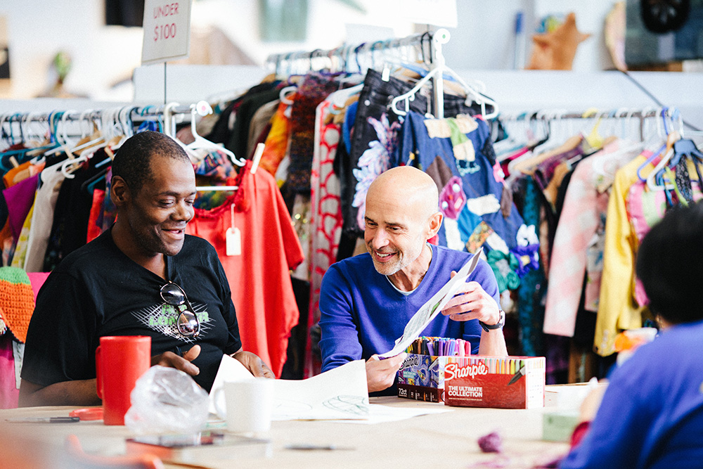 Two people sit at a table smiling surrounded by racks of colorful clothing.